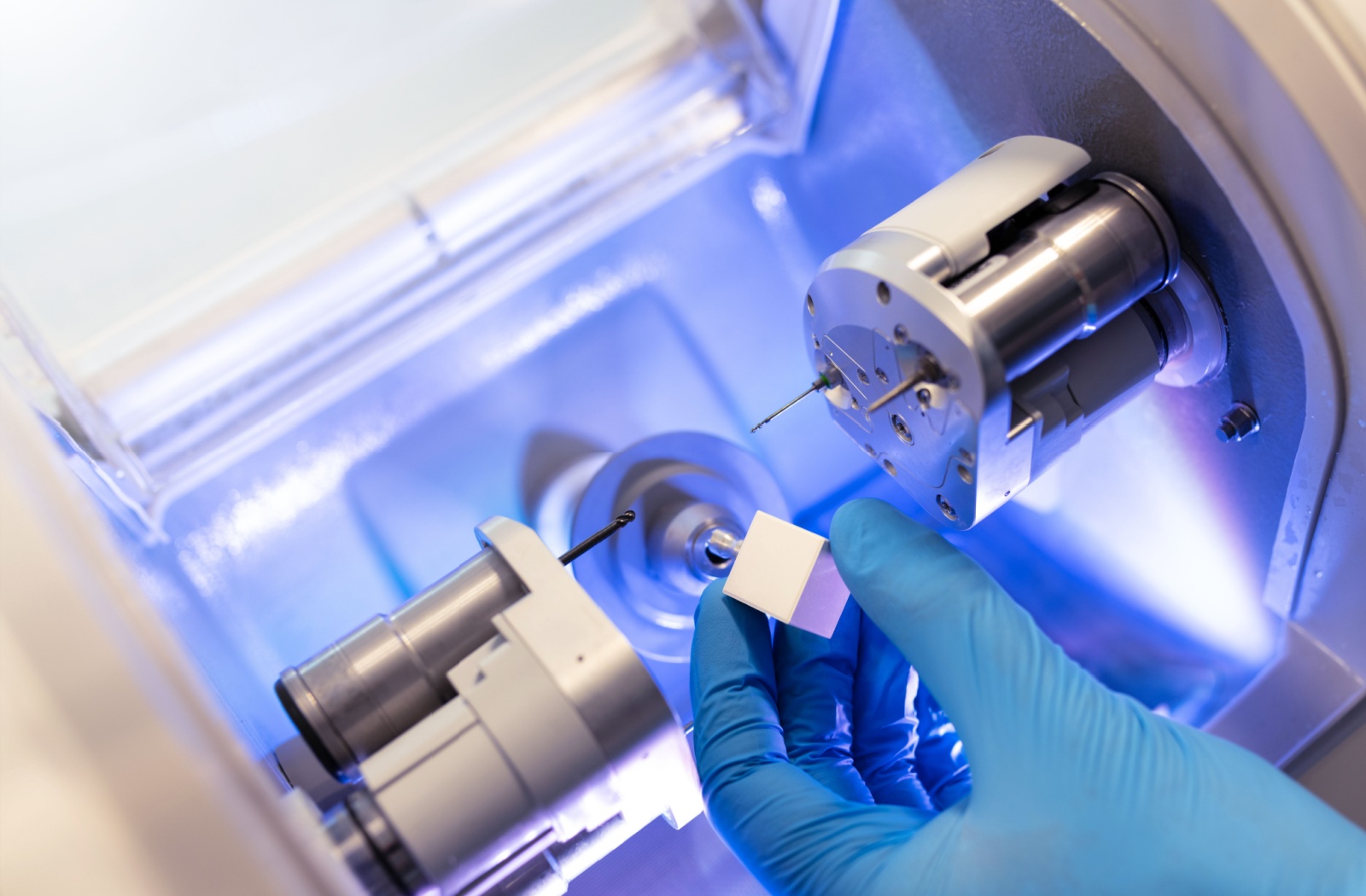 Close up of a person's hands in blue gloves holding a cube of material in a machine.