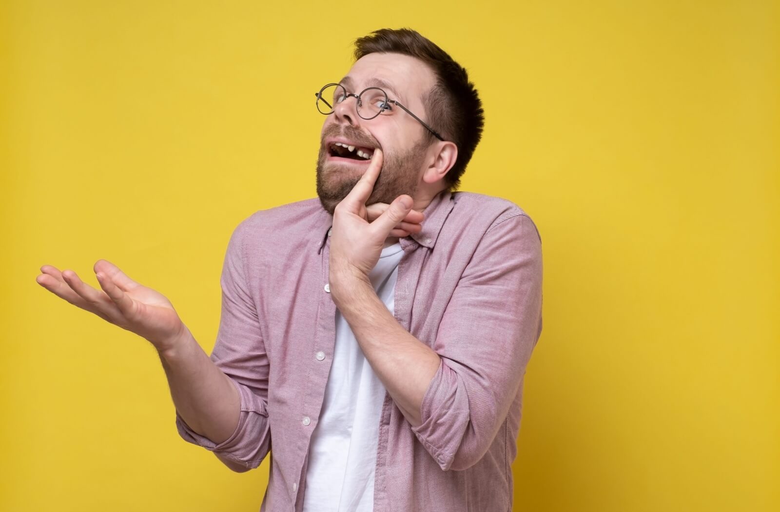 Man in glasses against a yellow background pulling back lip to reveal missing tooth. Other hand raised in perplexed gesture and expression.