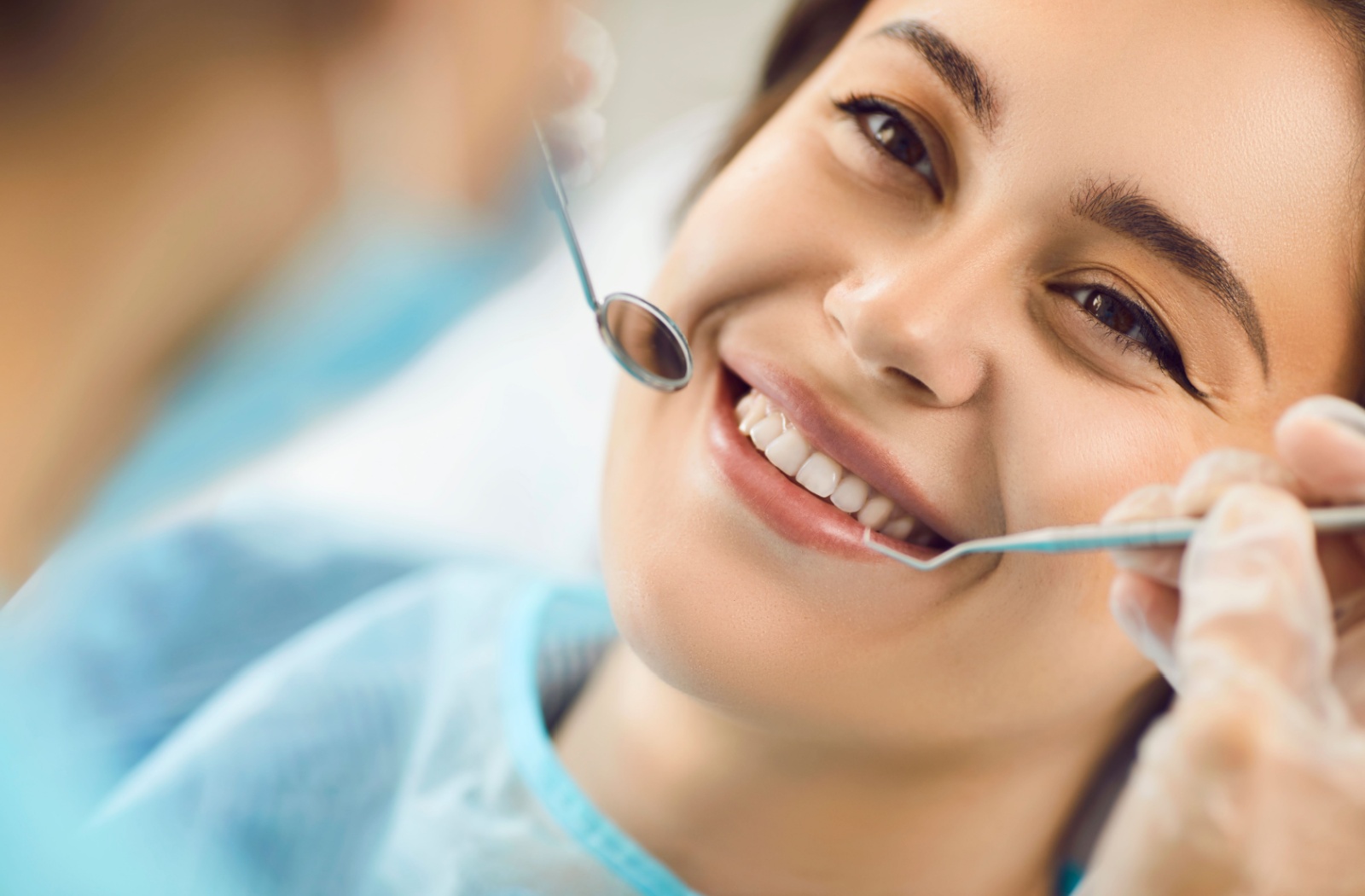 A dental patient smiles while a dentist uses a small mirror and pick to check their teeth.